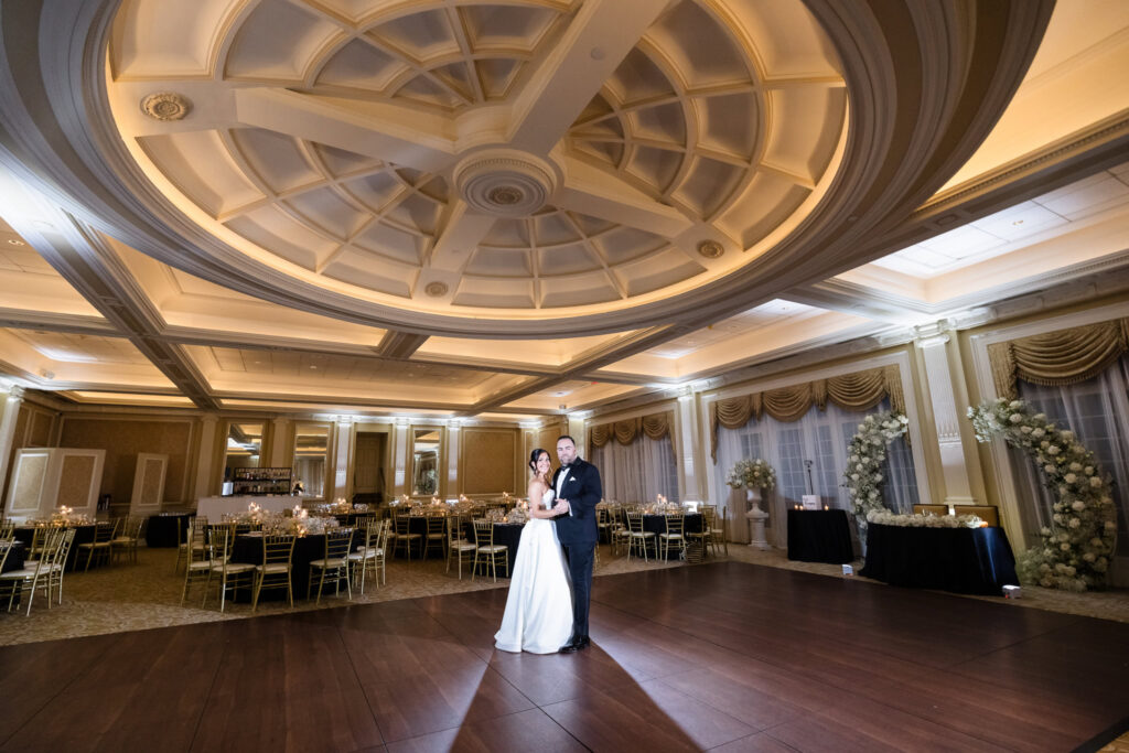 Bride and groom standing together on the dance floor beneath the grand illuminated ceiling of The Muttontown Club ballroom.