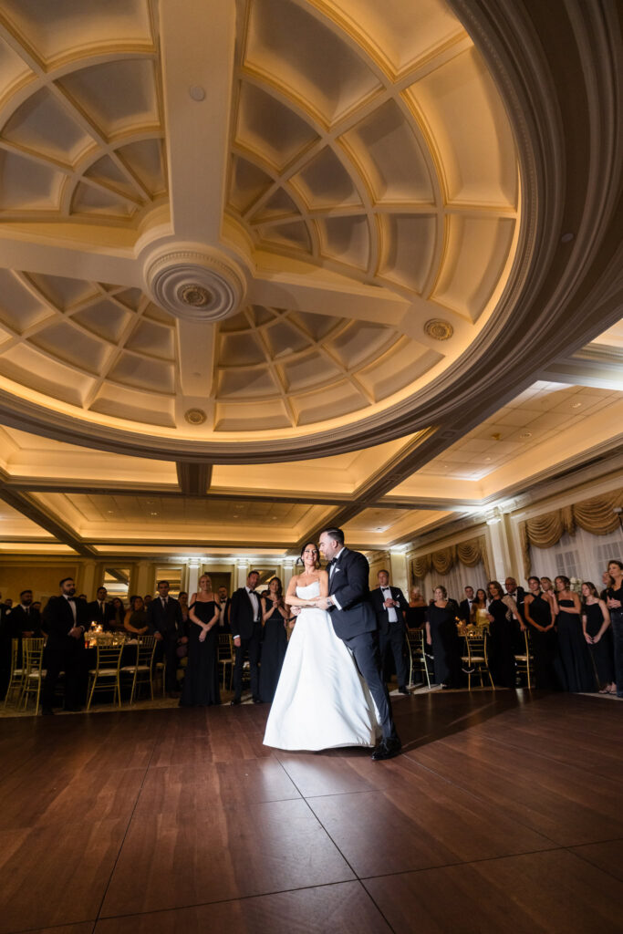 Bride and groom sharing their first dance surrounded by guests beneath the grand ballroom ceiling.