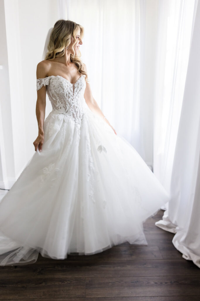 Kaity smiling beside a bright window in her off-the-shoulder wedding gown at Flowerfield.