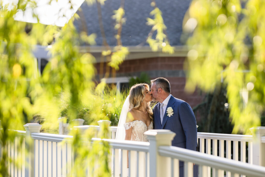 Kaity and Sean sharing a kiss on a white bridge surrounded by greenery during their summer wedding at Flowerfield on July 4th.