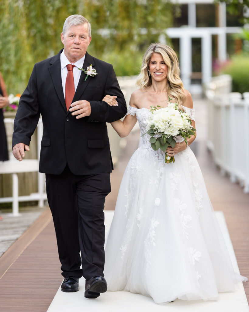 Kaity walking down the aisle with her father before her outdoor wedding ceremony at Flowerfield.
