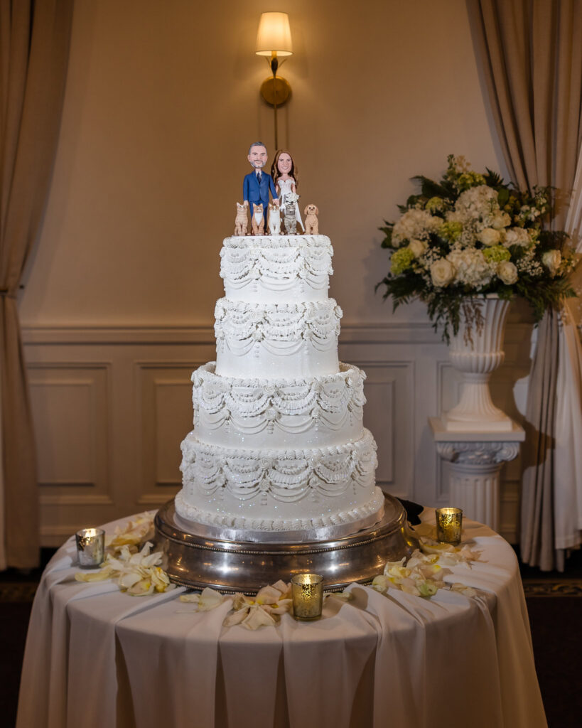 Four-tier white wedding cake with an ornate piped design and a custom topper featuring Kaity, Sean, and their pets on July 4th wedding.