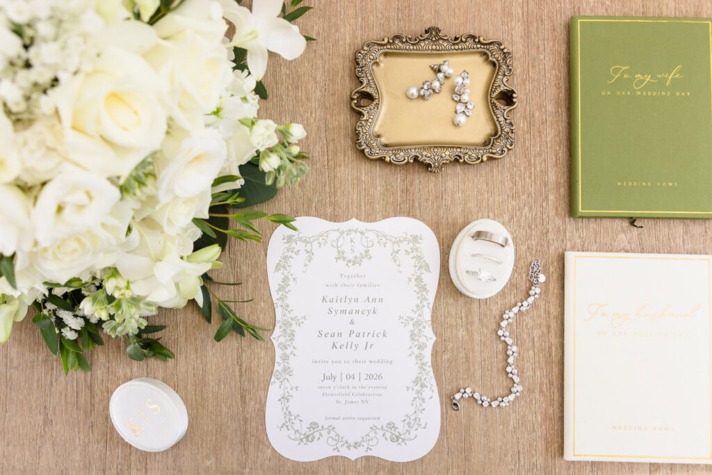 Kaity and Sean’s July 4th wedding invitation with white flowers, rings, jewelry, and wedding vow books.