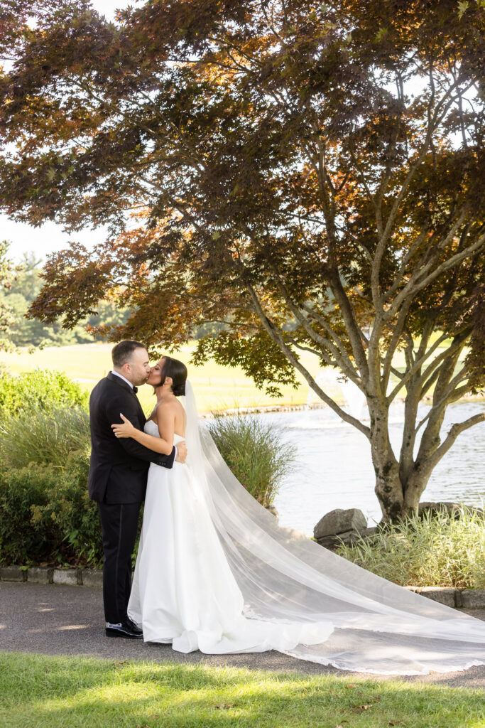 Bride and groom sharing a kiss beside the water beneath a tree, with the bride's long veil flowing behind her.