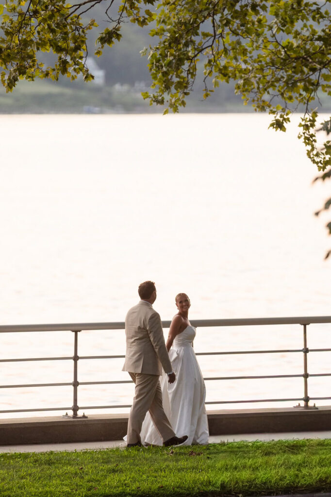 Bride and groom walking beside the waterfront at Sea Cliff Manor, framed beneath tree branches with Hempstead Harbor in the background.