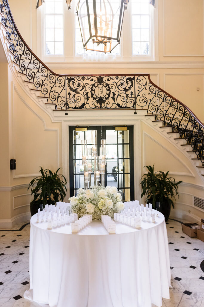 White floral arrangements, candles, and escort cards displayed beneath the grand staircase at The Muttontown Club.