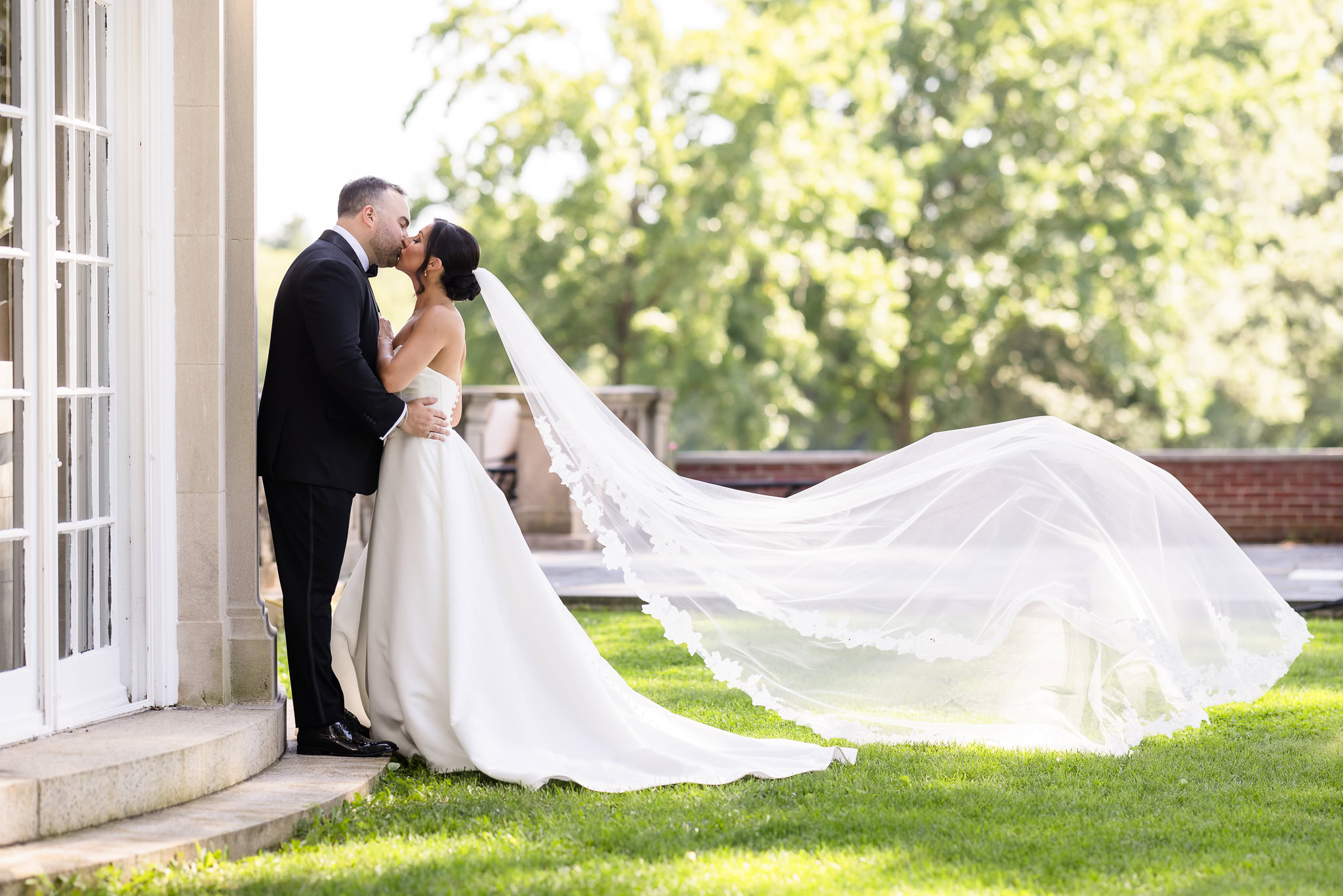 Christine and Matt kiss outdoors as her long veil catches the breeze during their Muttontown Club wedding portraits.