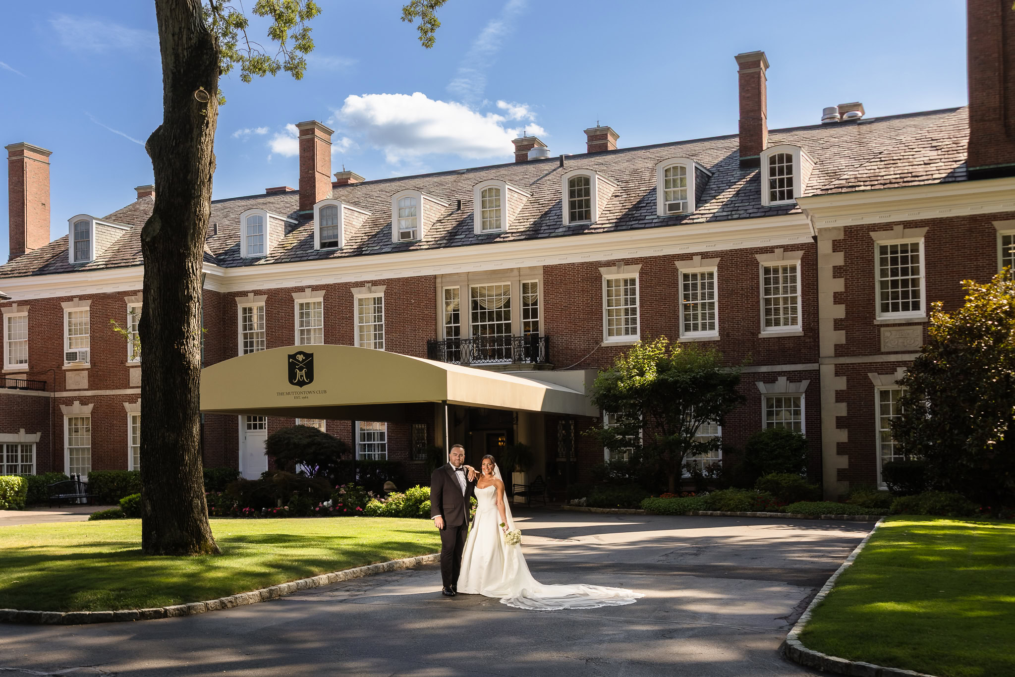 Christine and Matt stand together in front of the historic brick clubhouse during their Muttontown Club wedding.