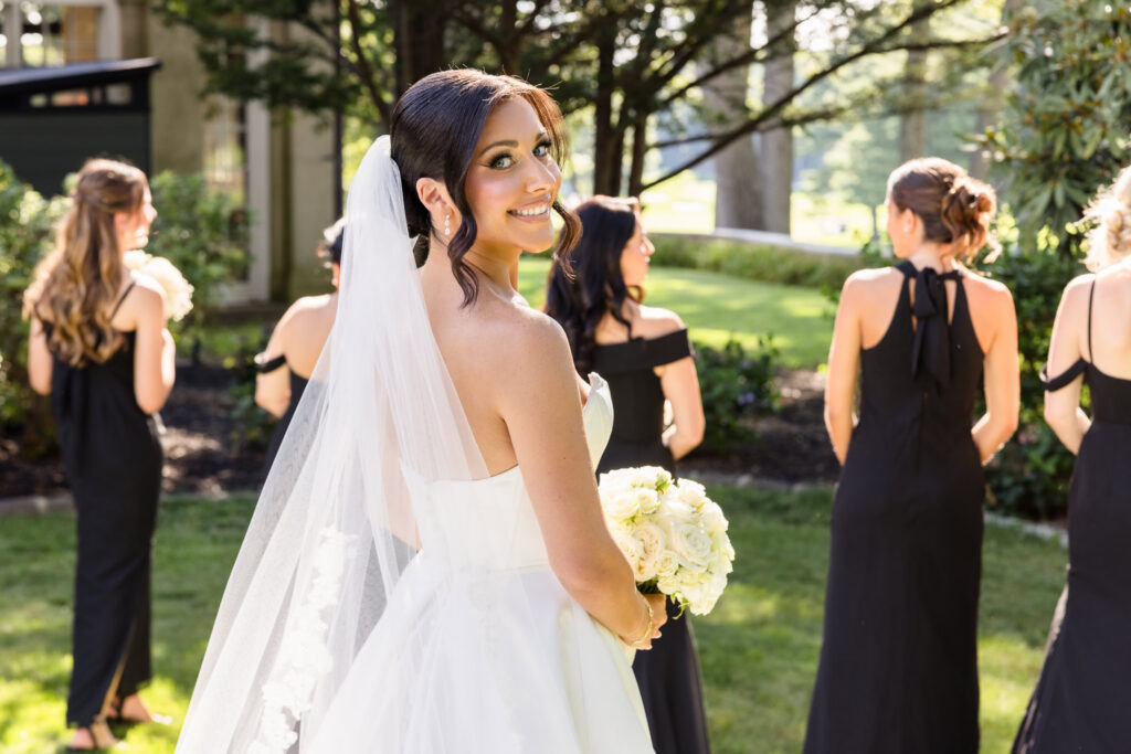 Christine smiles toward the camera while holding a white rose bouquet with her bridesmaids nearby on the grounds.