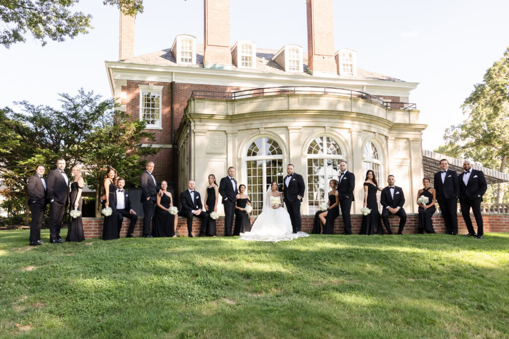 Christine and Matt pose with their large wedding party in black formal attire outside The Muttontown Club.
