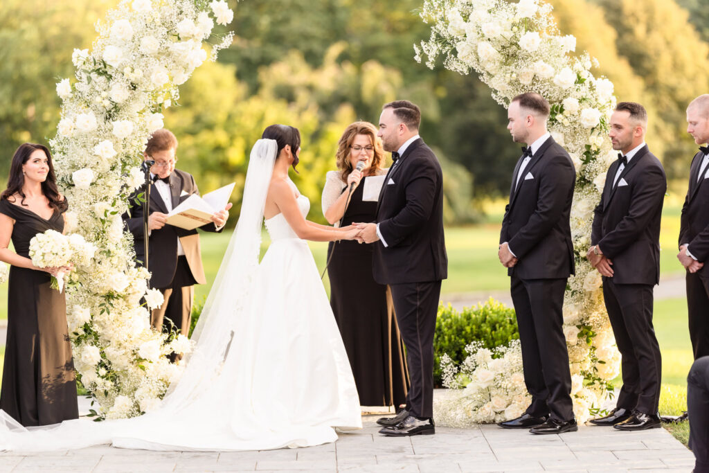 Bride and groom exchanging vows beneath white floral arches during an outdoor Muttontown Club wedding ceremony.