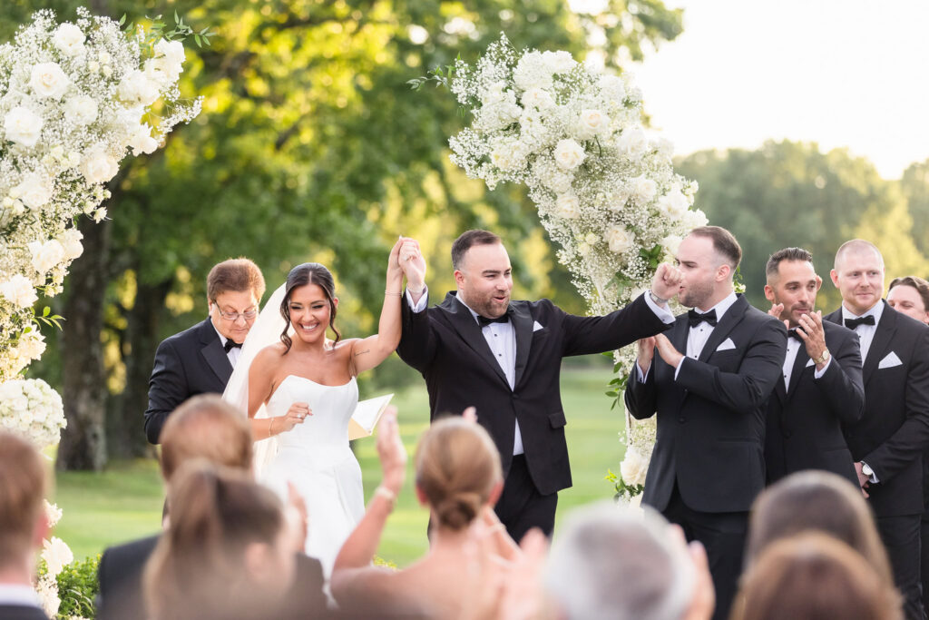 Newlyweds celebrating after their vows beneath white floral arrangements at The Muttontown Club.