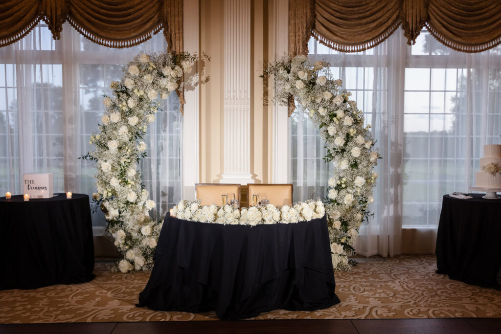 Black-draped sweetheart table framed by white floral installations inside The Muttontown Club ballroom.