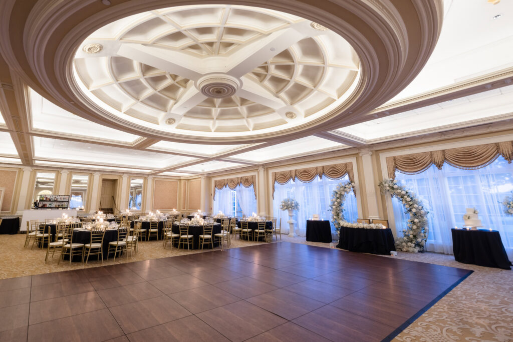 Wide view of The Muttontown Club ballroom prepared for a wedding reception with black linens and gold Chiavari chairs.