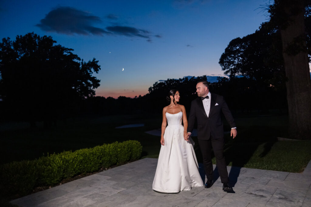 Bride and groom walking together beneath a crescent moon at twilight.