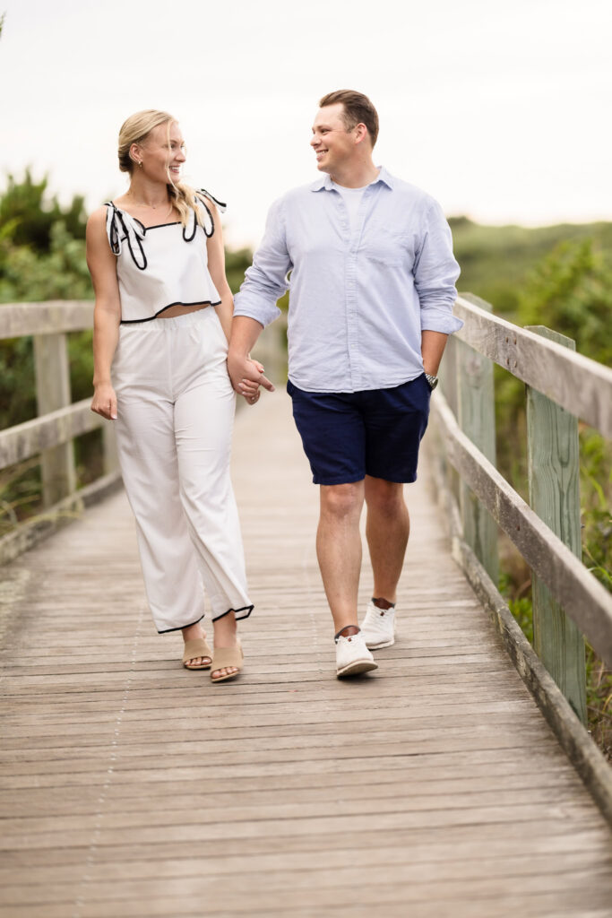 Emily and Corbin hold hands and smile while walking along the boardwalk during their Robert Moses engagement session.