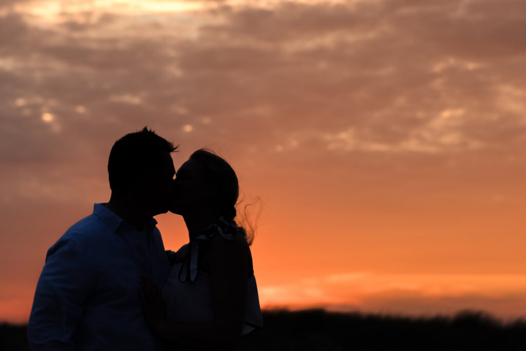 Emily and Corbin kiss in silhouette beneath a dramatic orange sunset at Robert Moses.