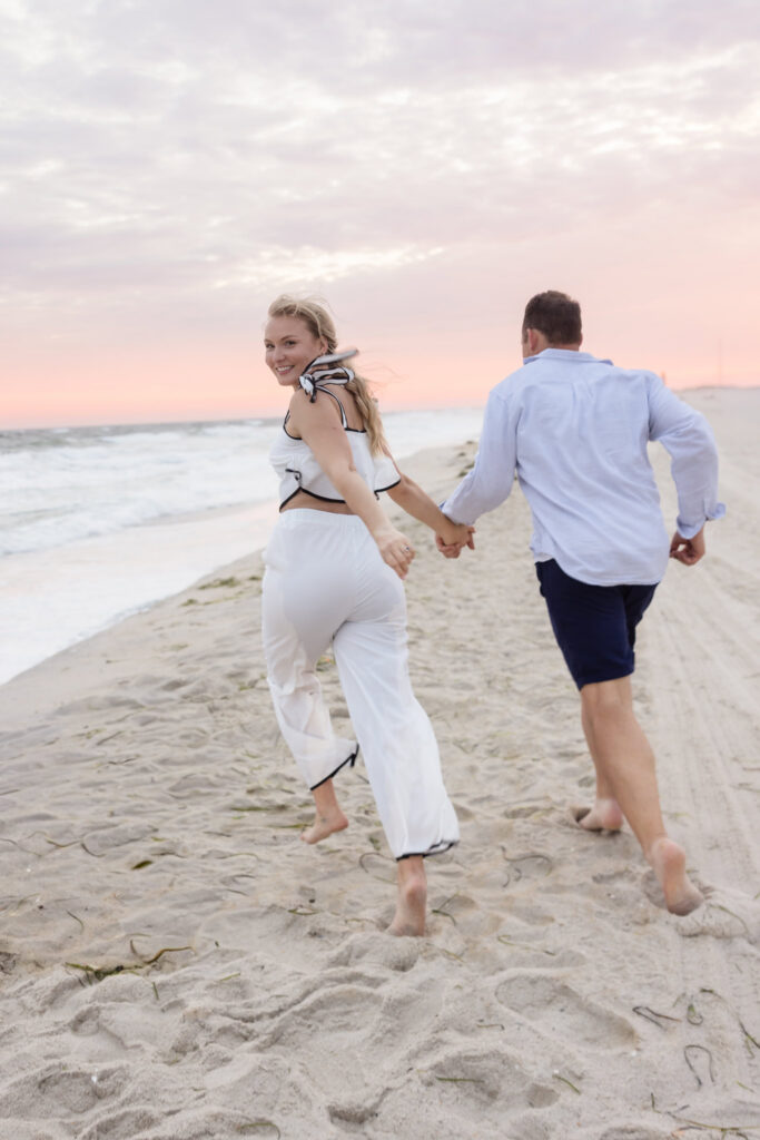 Emily looks back smiling as she and Corbin run barefoot along the beach during their engagement session.