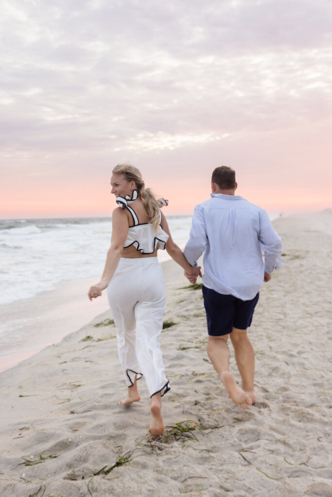 Emily and Corbin run hand in hand along the shoreline beneath a soft pink sky at Robert Moses.