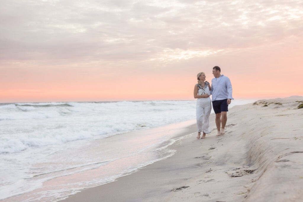 Emily and Corbin walk together beside the ocean under a pastel sunset during their Long Island engagement session.