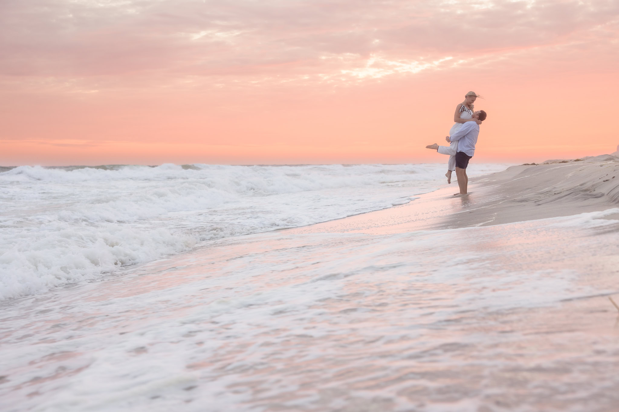 Emily and Corbin embrace at the water's edge beneath a pink sunset during their Robert Moses engagement session.