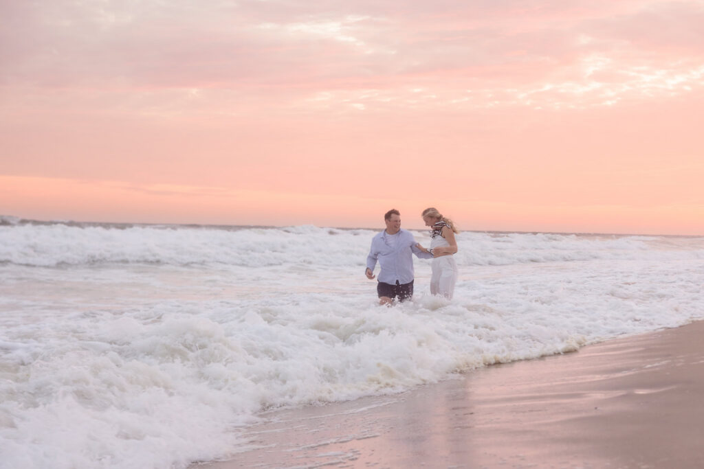 Emily and Corbin stand together in the waves beneath a glowing pink sky at Robert Moses.