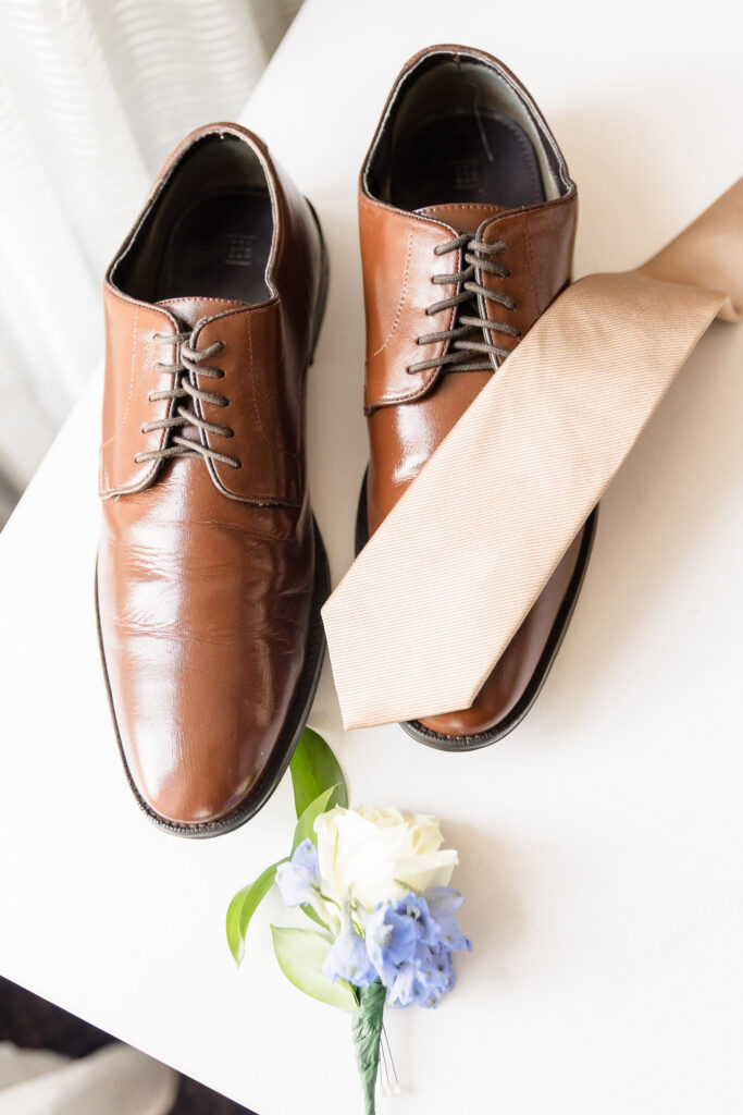 Groom’s brown leather shoes, neutral tie, and white and blue boutonniere arranged together before the wedding.