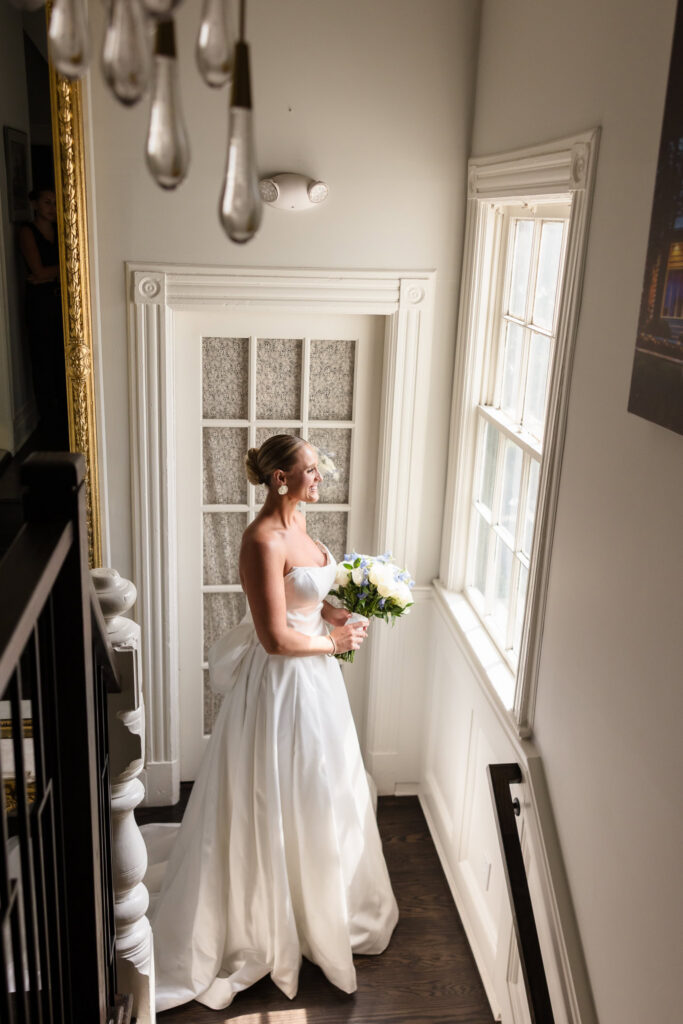 Bride standing by a sunlit window with her bouquet before her Sea Cliff Manor wedding.