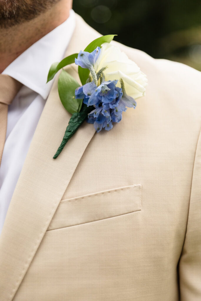 Close-up of Josh’s blue and white floral boutonniere on his tan wedding suit.