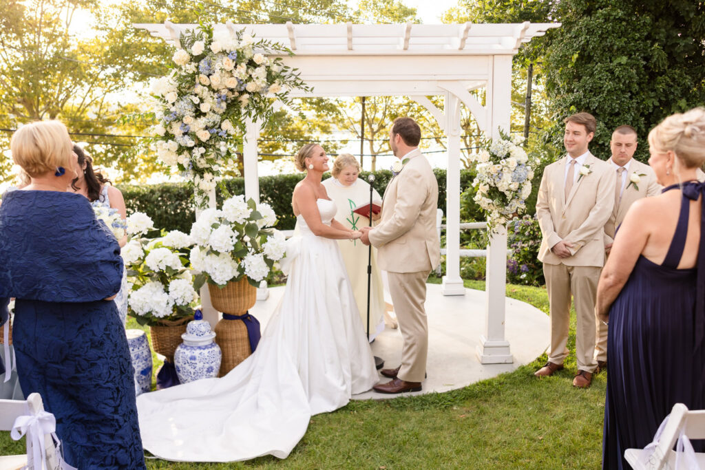 Quinn and Josh exchange vows beneath a white floral arbor during their large bridal party wedding ceremony at Sea Cliff Manor.