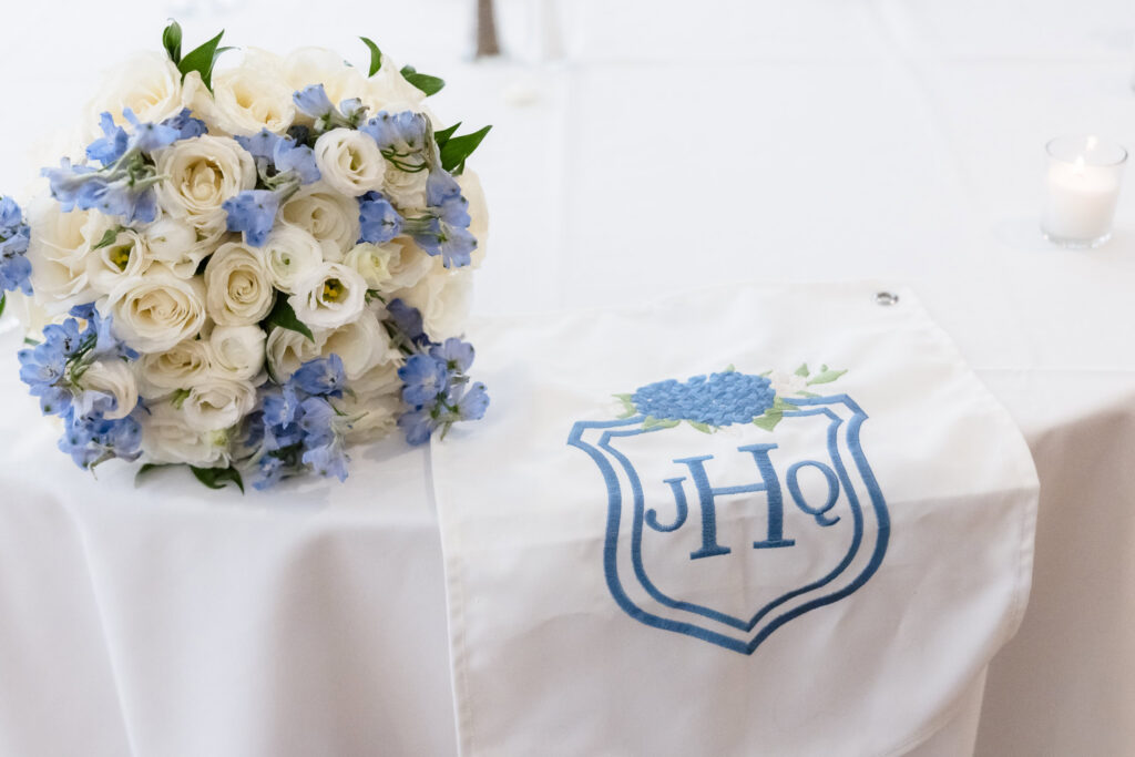 White roses and blue flowers beside a custom embroidered monogram at Quinn and Josh’s Sea Cliff Manor wedding.