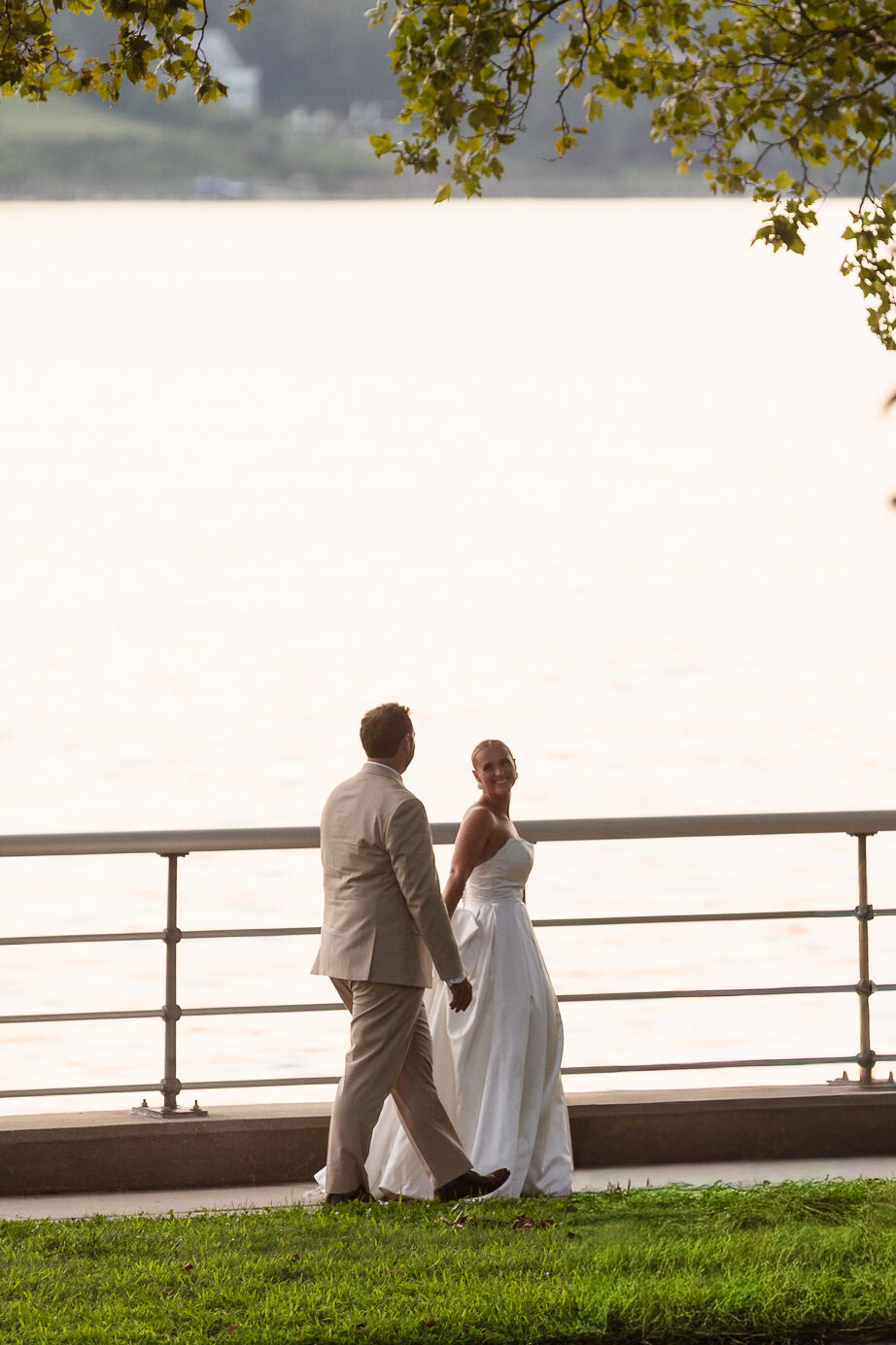 Quinn and Josh walk together beneath the trees overlooking the water at sunset near Sea Cliff Manor.