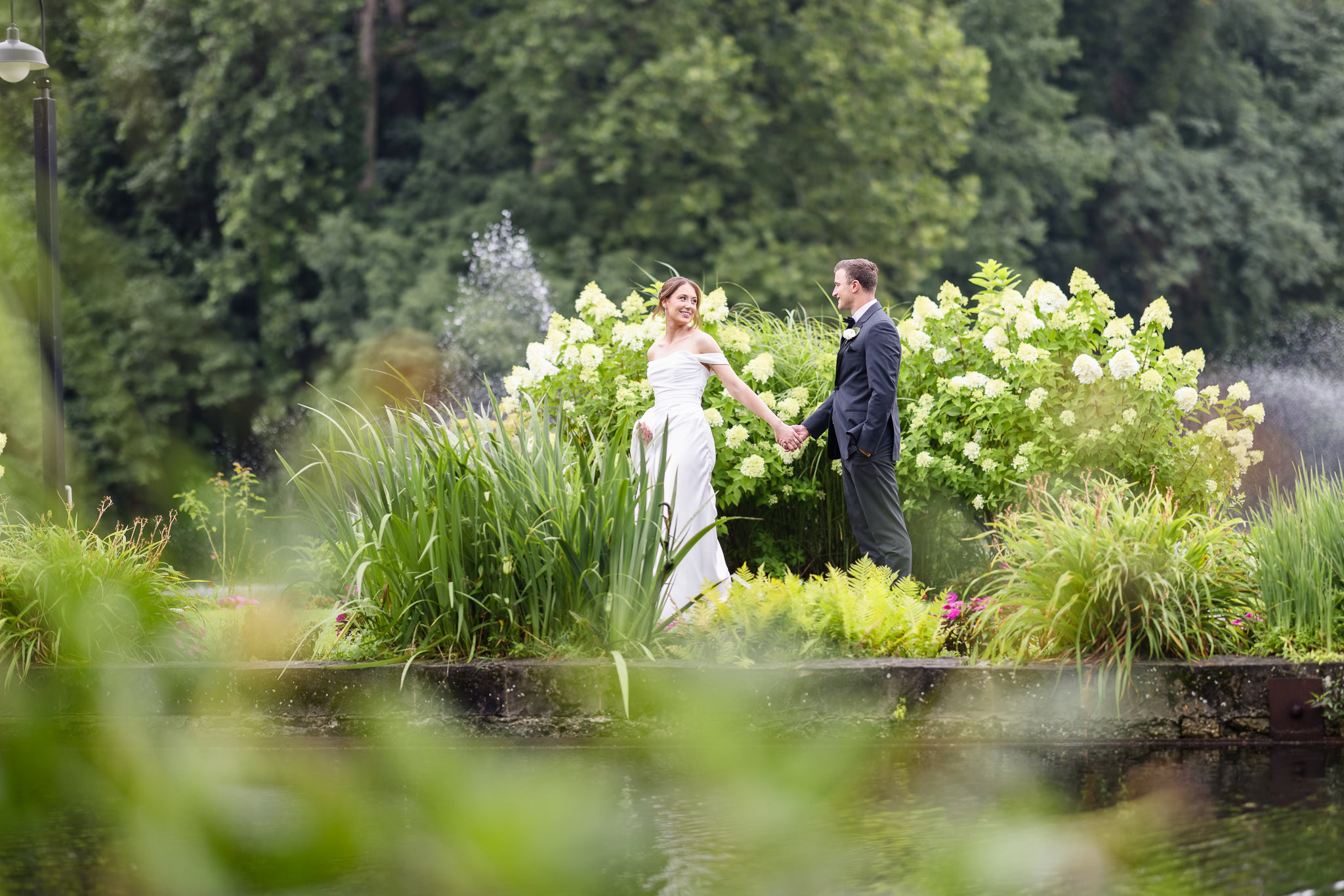 Bride and groom hold hands beside the gardens and water during their Swan Club wedding portraits.