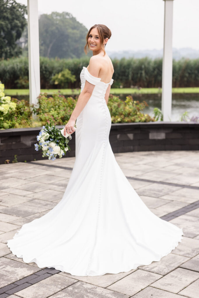 Bride smiling over her shoulder while showing the train and button details of her off-the-shoulder wedding dress.