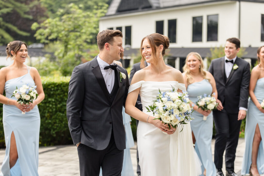 Bride and groom smiling together with bridesmaids in light blue dresses and groomsmen in black tuxedos behind them.