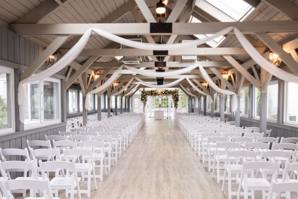 Bright indoor wedding ceremony space with rows of white chairs, exposed beams, white draping, and a floral ceremony arch.