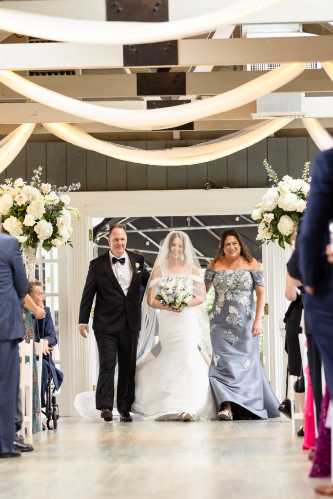 Bride walking down the wedding ceremony aisle with her parents while carrying a white and blue bouquet.