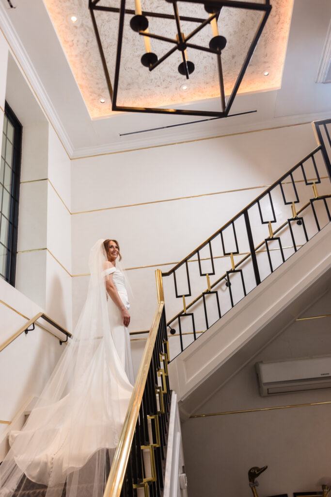 Bride in an off-the-shoulder wedding gown and long veil posing on the elegant staircase at The Swan Club