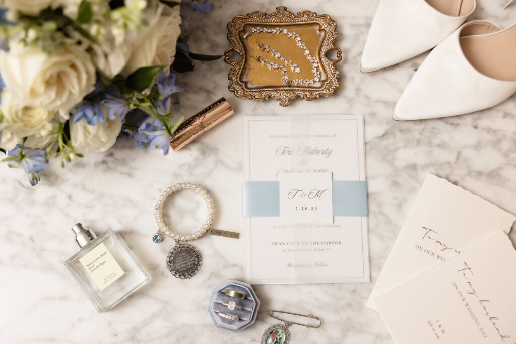 Wedding invitation, jewelry, white heels, perfume, rings, and blue-and-white florals arranged for a Swan Club on the Harbor wedding.