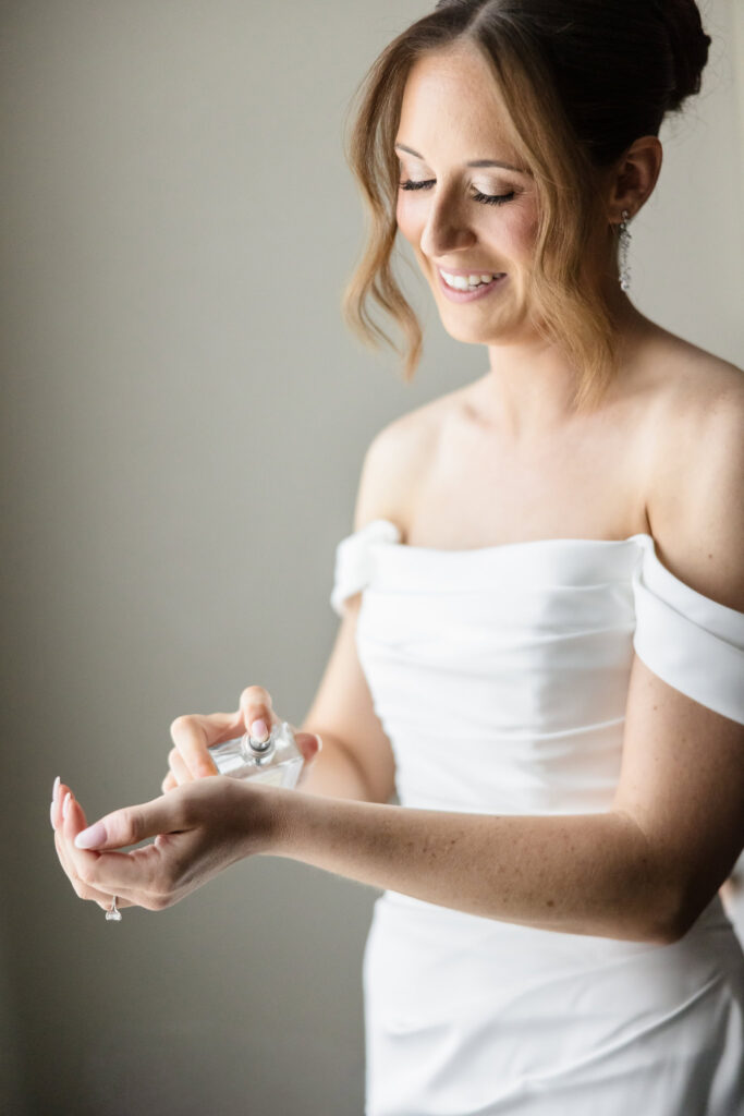Bride in an off-the-shoulder wedding gown applying perfume while getting ready.