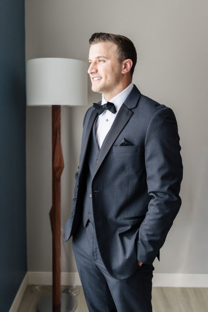 Groom in a charcoal tuxedo and black bow tie standing beside a window before the wedding.