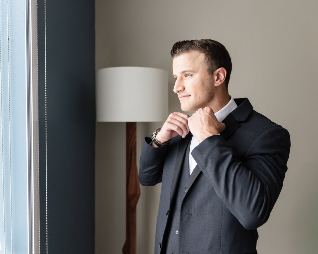 Groom adjusting his black bow tie by the window while preparing for the wedding day.