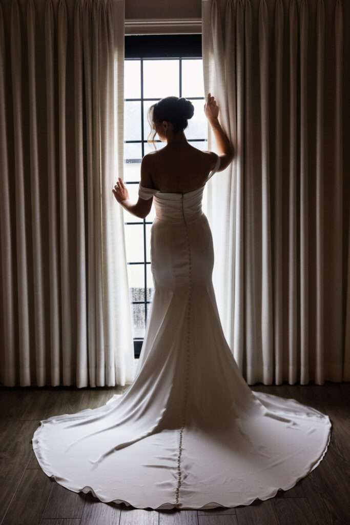 Bride standing in front of a tall window, showing the elegant silhouette and train of her wedding gown.