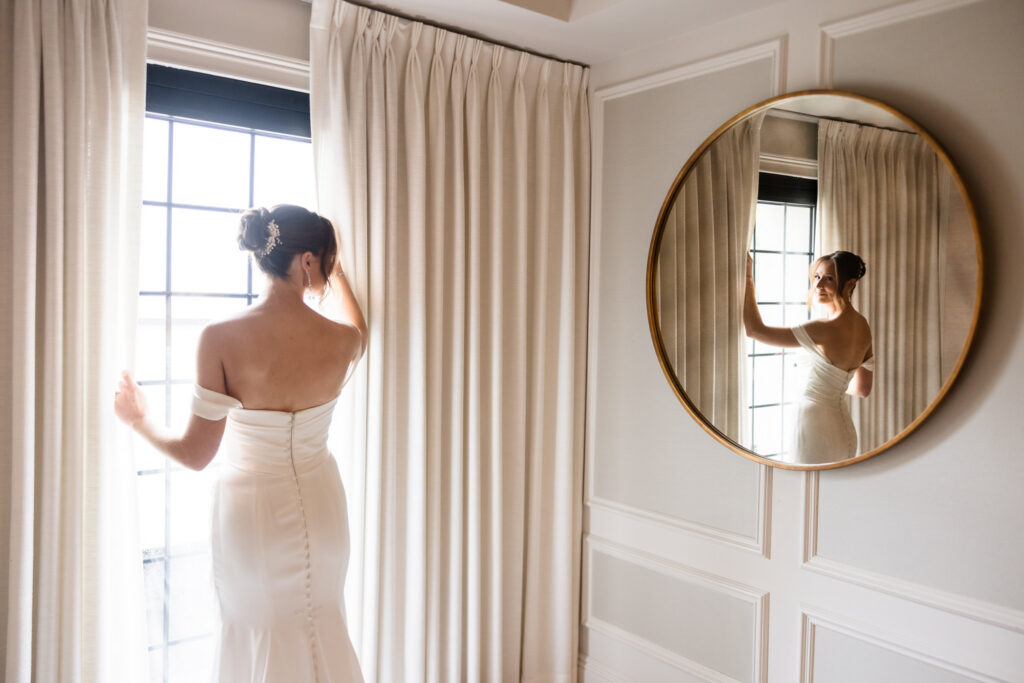 Bride standing beside a bright window with her reflection framed in a round gold mirror.