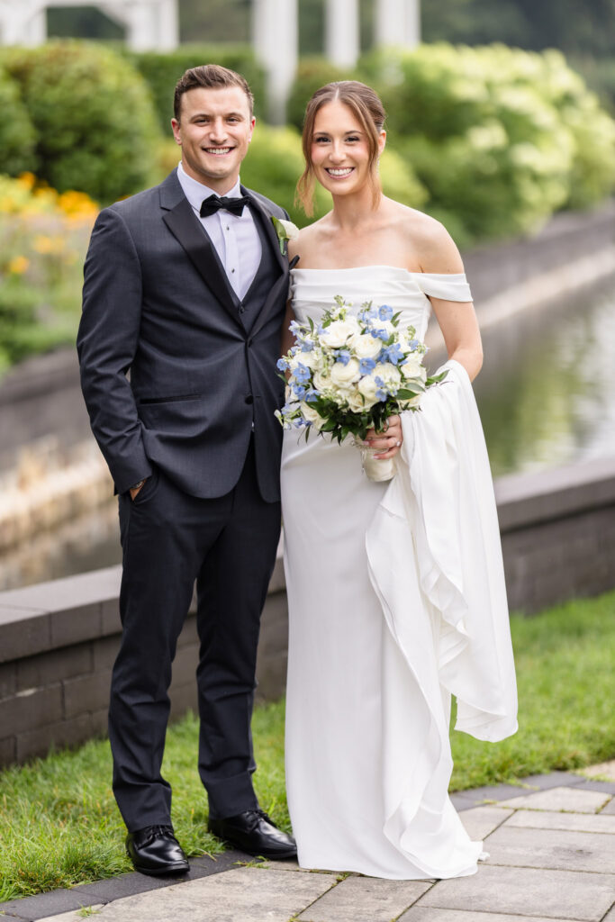 Bride and groom smiling together with a blue-and-white bouquet beside the venue gardens.