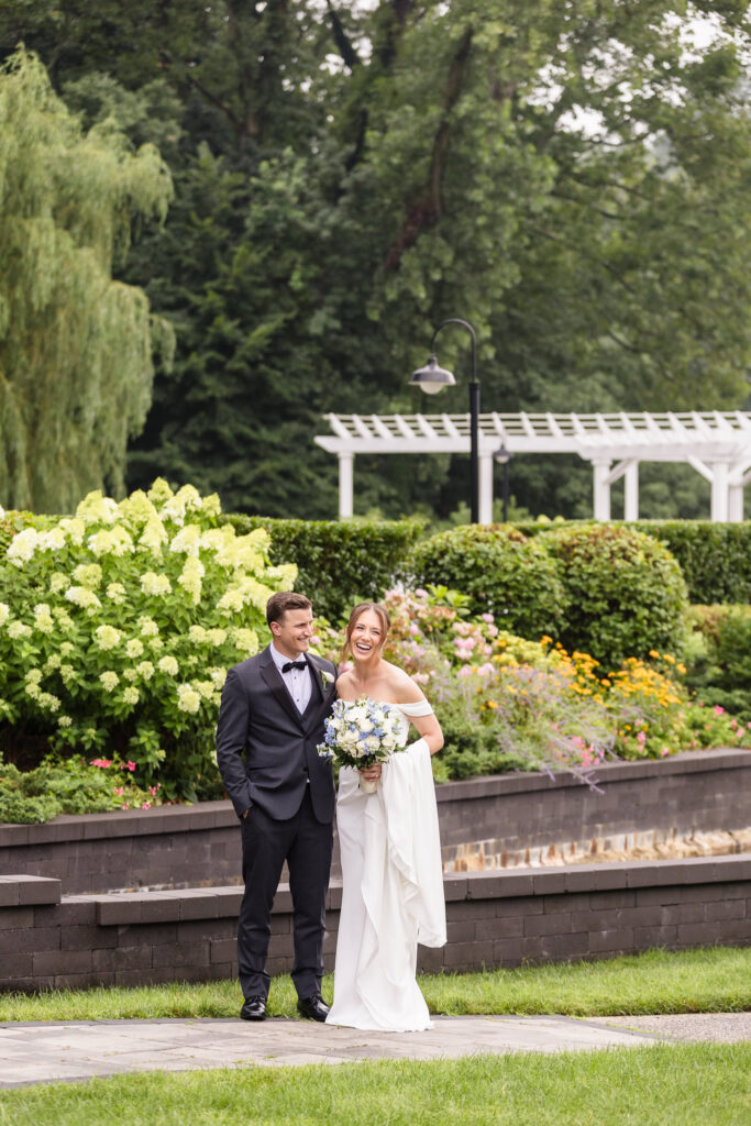 Bride and groom laughing together among lush hydrangeas and colorful summer flowers.