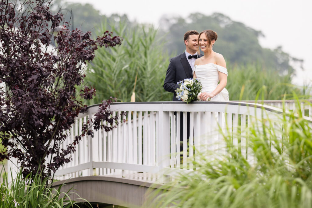 Bride and groom smiling together on a white garden bridge surrounded by lush greenery.