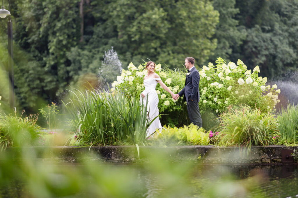 Bride and groom holding hands across a garden pond surrounded by greenery and blooming hydrangeas.