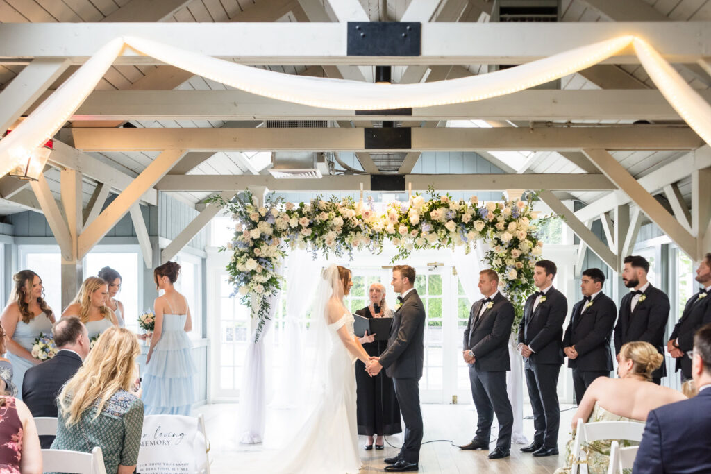 Tori and Mark hold hands beneath a floral arch during their Swan Club pavilion wedding ceremony.