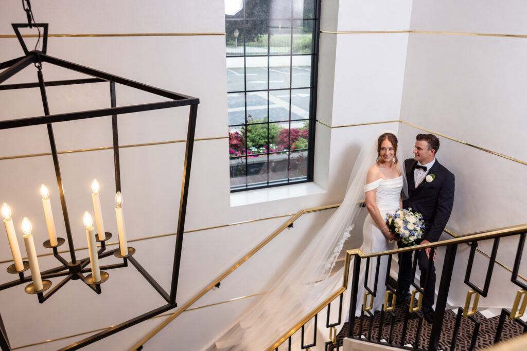 Tori and Mark pose together on the staircase inside The Swan Club beneath a modern chandelier.